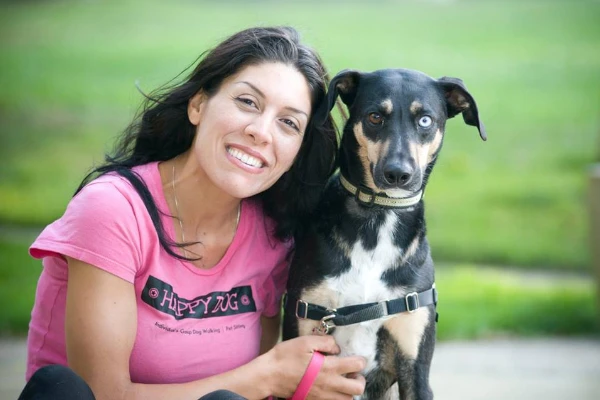 A woman in a pink shirt smiles while sitting next to a black and white dog with blue and brown eyes. They are outdoors with a grassy background.