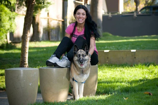 A woman in a pink shirt and black leggings sits on a stone stool at a park, smiling. A large dog with a harness stands beside her.