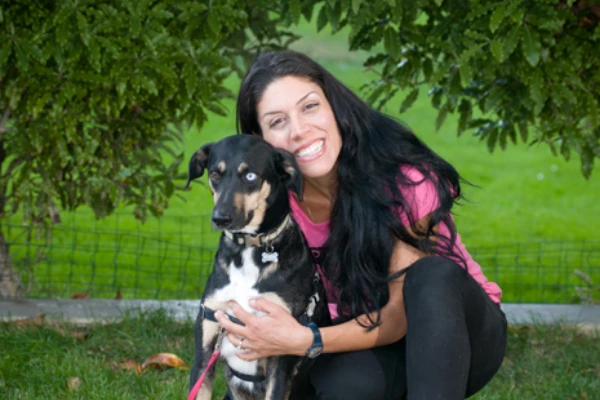 A woman in a pink shirt and black pants smiles while holding a black and tan dog with blue eyes in front of green foliage.