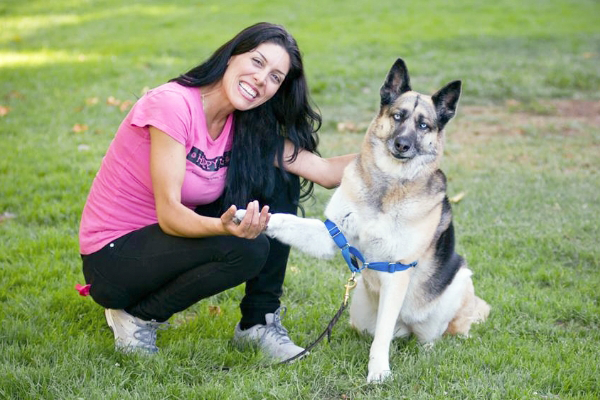 A woman with long dark hair wearing a pink shirt smiles while shaking hands with a large dog in a blue harness, perfectly capturing a moment for the Happy Dog Gallery.