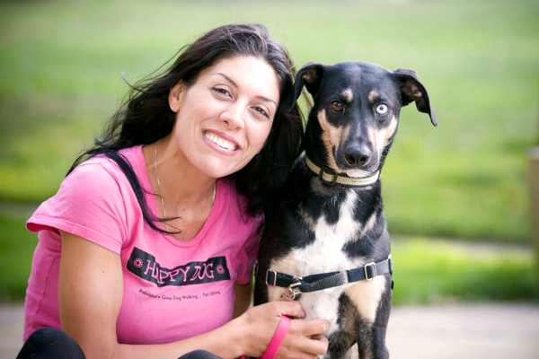 A woman in a pink shirt smiles while sitting next to a black dog with blue and brown eyes. The woman is holding the dog's leash, creating a perfect moment for the Happy Dog Gallery. They are outdoors on a grassy area.