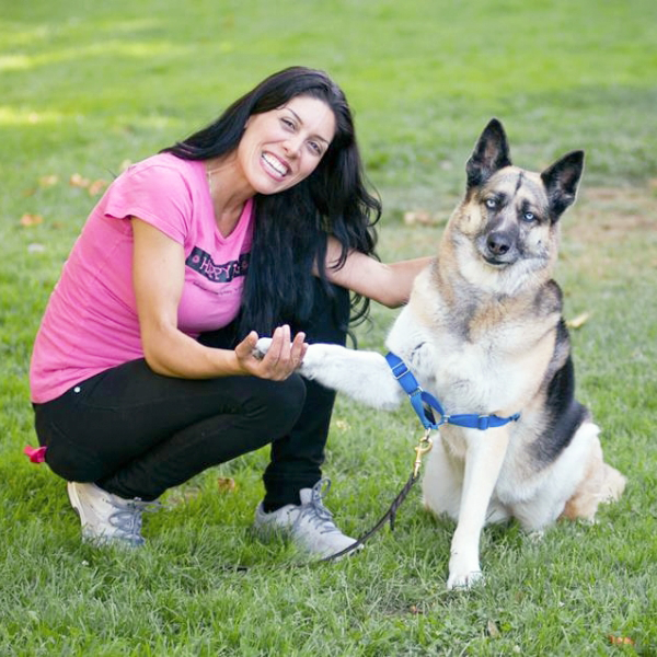 A dog walker on the grass shakes hands with a large dog on a leash. Stefania is smiling and wearing a pink shirt, connects with the light brown and black-coated canine in a heartwarming moment.